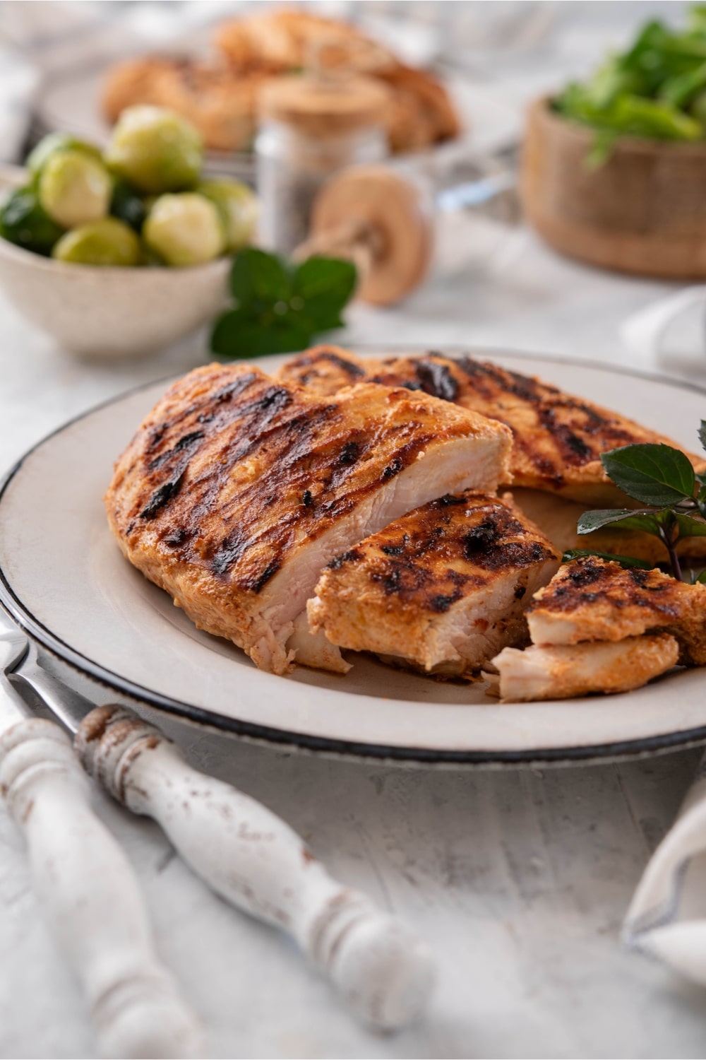 Sliced grilled chicken breasts on a white plate, surrounded by bowls of salad greens and brussels sprouts.