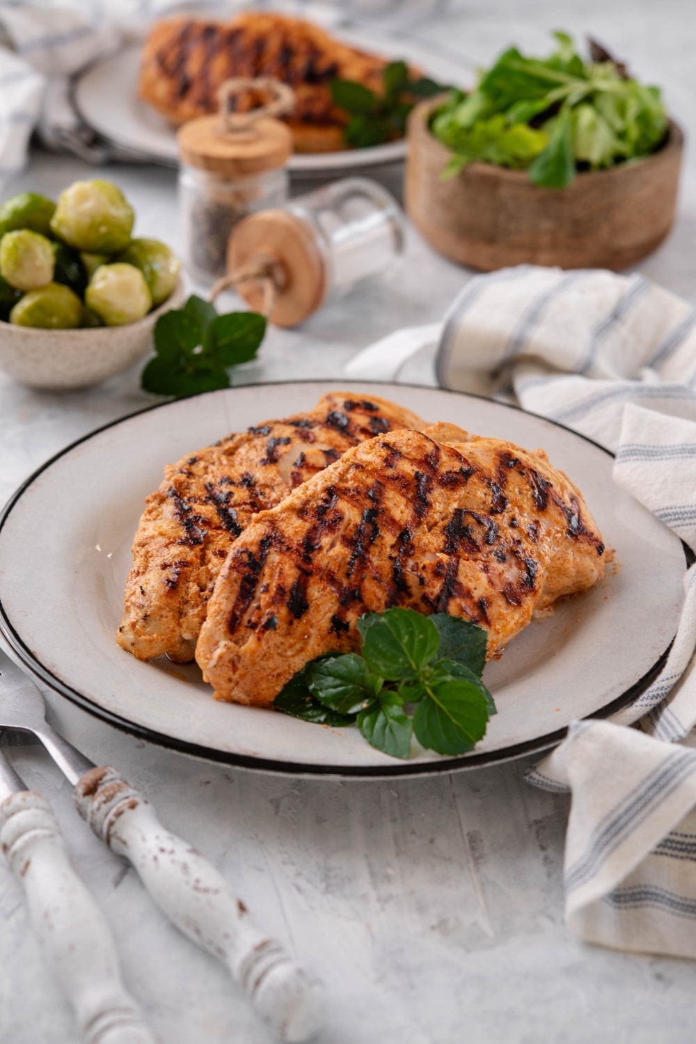 Grilled yogurt-marinated chicken breasts on a white plate, surrounded by bowls of salad greens and brussels sprouts.