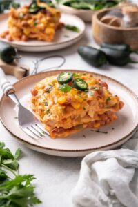 A slice of enchilada casserole with melted cheese, corn, chicken, beans, and peppers in it. The casserole is garnished with two jalapeño slices and fresh herbs and there is a fork on the plate.