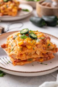 A serving of enchilada casserole with melted cheese, corn, chicken, beans, and peppers in it. The casserole is garnished with two jalapeño slices and fresh herbs and there is a fork on the plate.