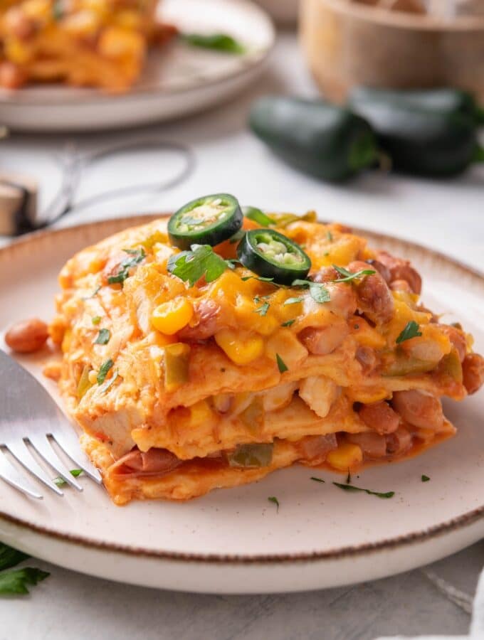 A serving of enchilada casserole with melted cheese, corn, chicken, beans, and peppers in it. The casserole is garnished with two jalapeño slices and fresh herbs and there is a fork on the plate.