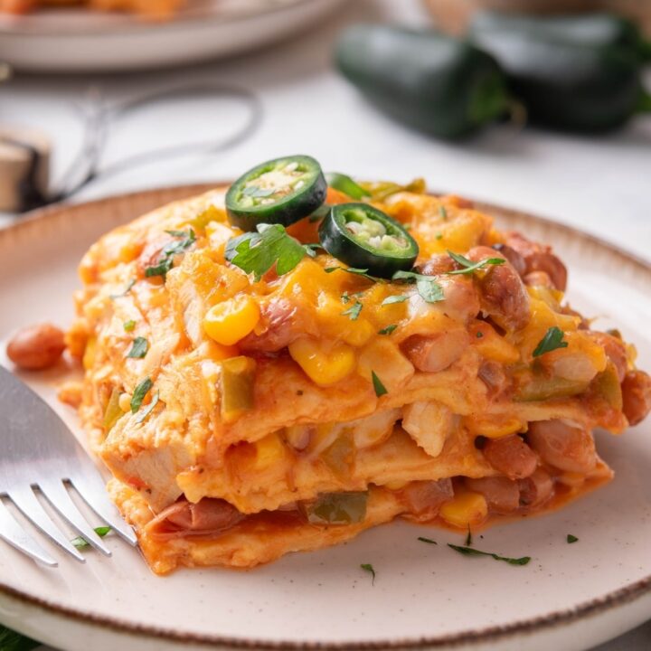 A serving of enchilada casserole with melted cheese, corn, chicken, beans, and peppers in it. The casserole is garnished with two jalapeño slices and fresh herbs and there is a fork on the plate.