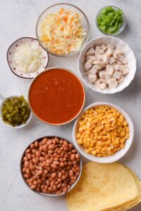 An assortment of ingredients including bowls of enchilada sauce, diced chicken, pinto beans, corn, shredded cheese, diced green peppers, diced onion, green chiles, and a stack of tortillas all on a white counter.