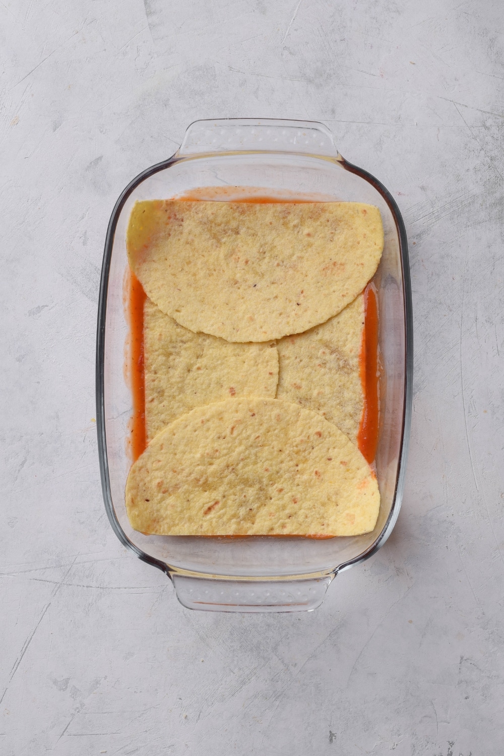 A baking dish with sauce and halved tortillas lining the bottom of the baking dish.