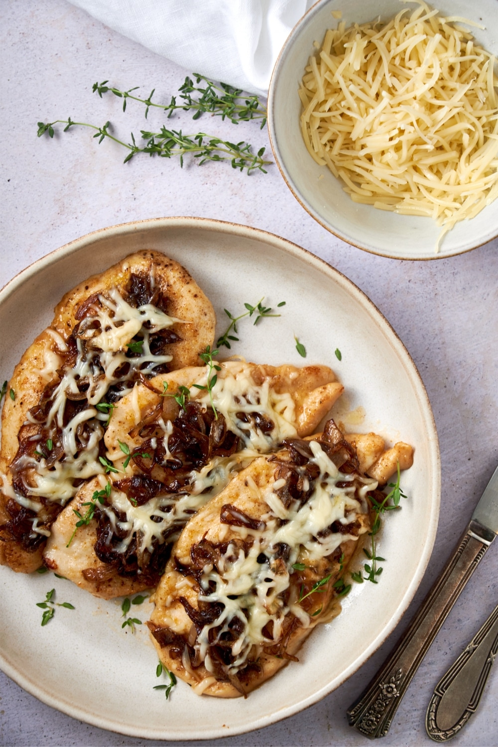 French onion chicken cutlets on a plate garnished with fresh thyme next to a bowl of shredded cheese.