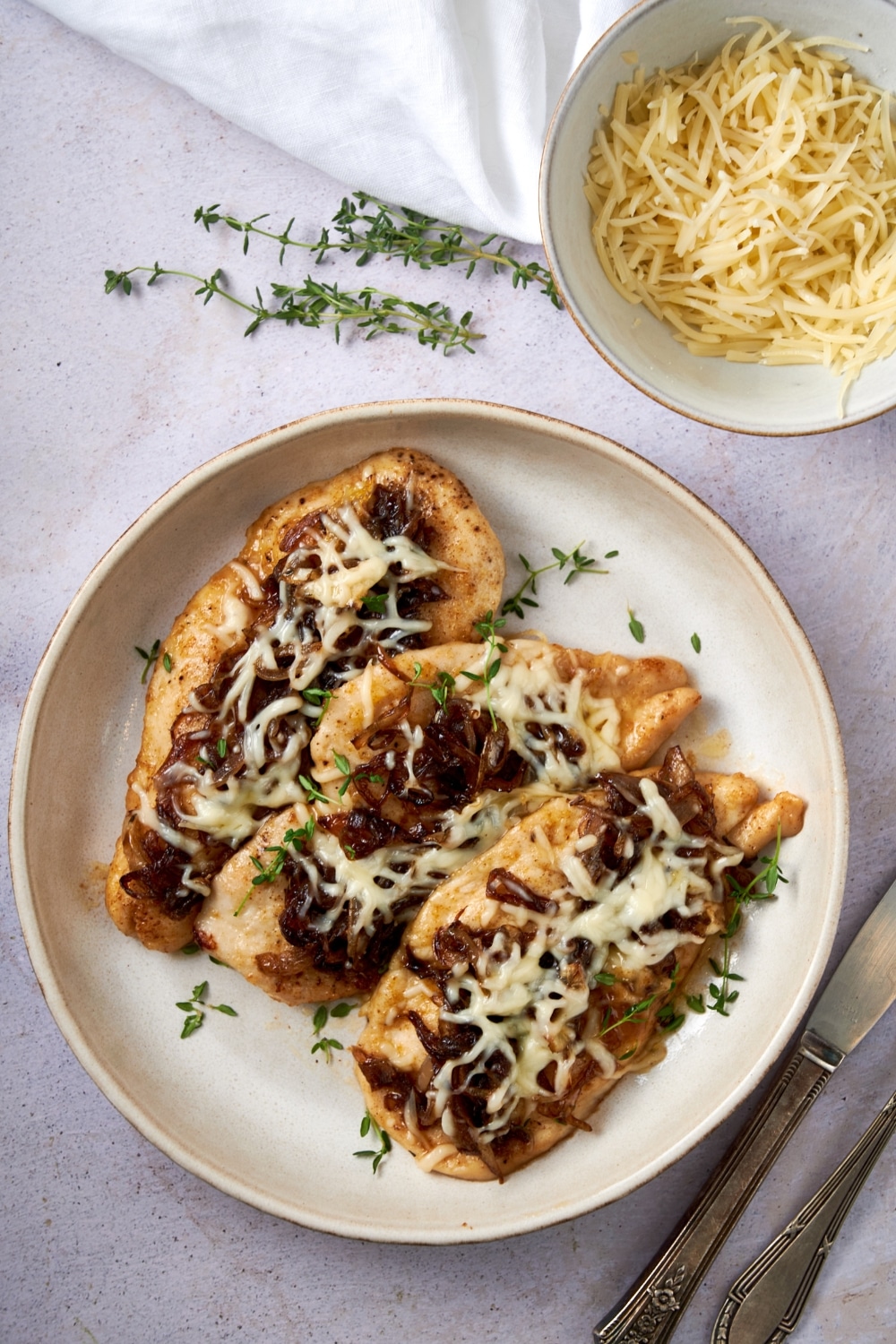 Three pieces of French onion chicken on a plate garnished with fresh thyme next to a bowl of shredded cheese.