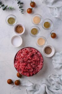 Ground beef in a bowl and eight bowls of different seasonings all on a white counter.
