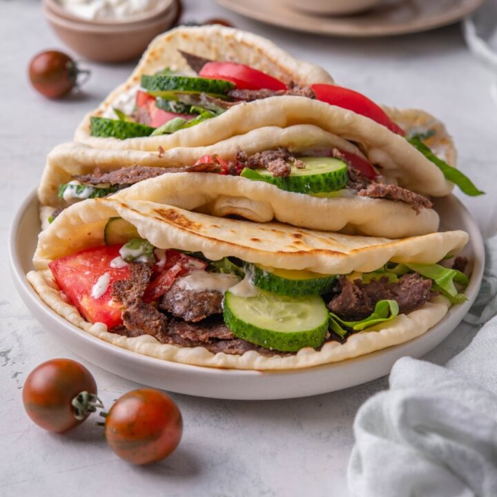 Three gyros piled on top of each other, each with thinly sliced meat, sliced cucumber, lettuce, tomato and tzatziki sauce.