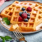 A fork with a piece of waffle on it in front of a plate with three waffles.