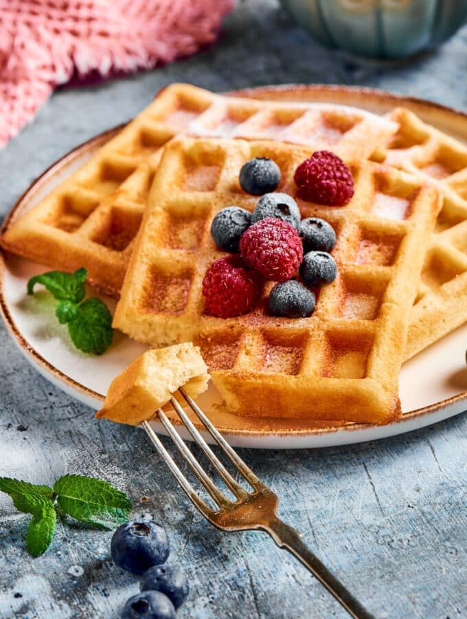 A fork with a piece of waffle on it in front of a plate with three waffles.