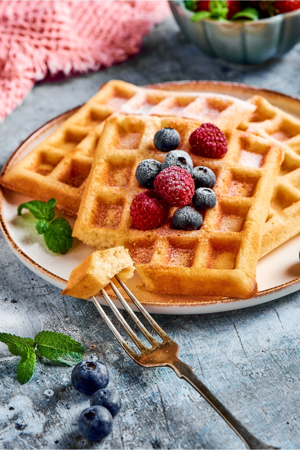 A fork with a piece of waffle on it in front of a plate with three waffles.