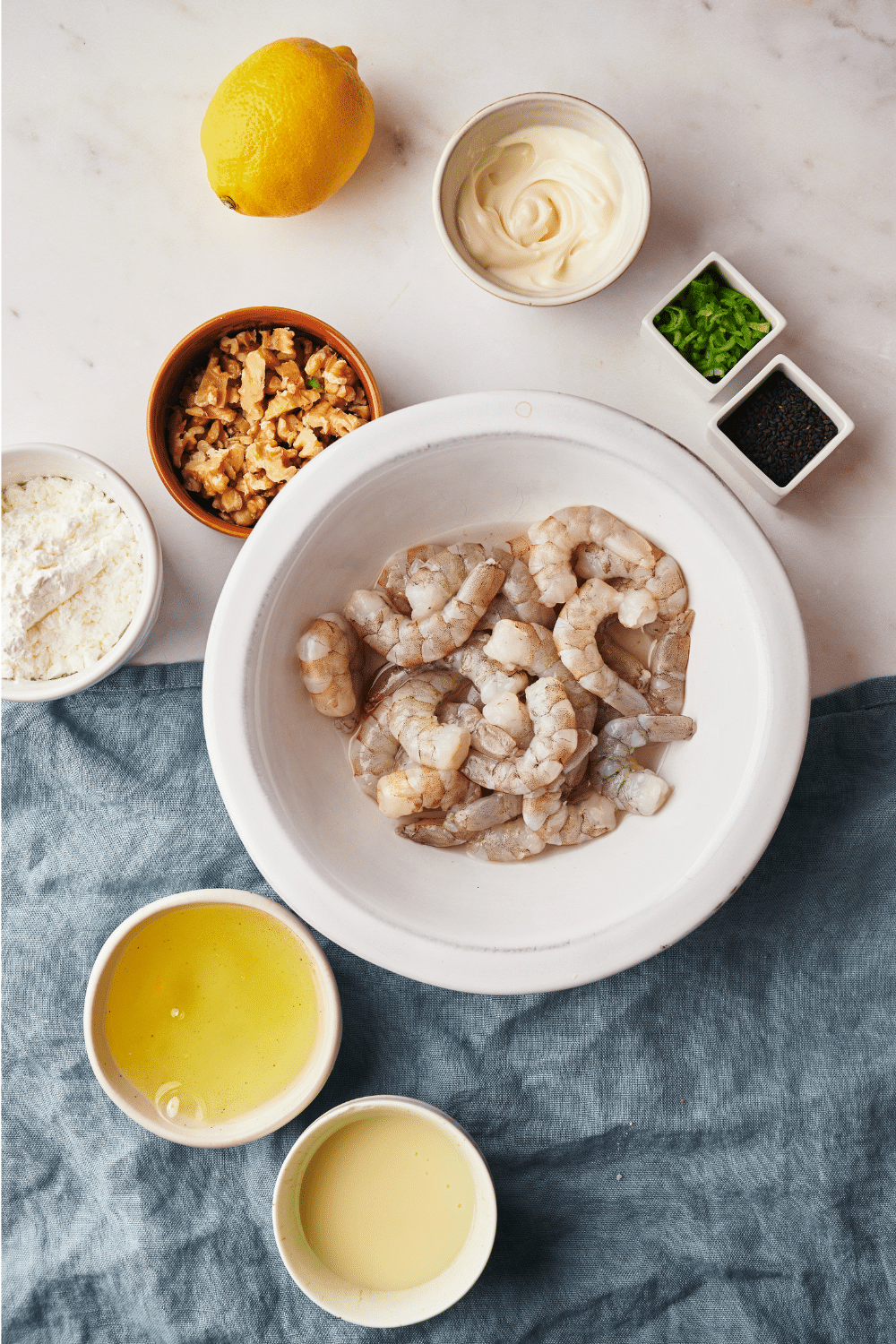 An assortment of ingredients including bowls of raw shrimp, walnuts, egg whites, mayonnaise, flour, green onions, and a lemon.