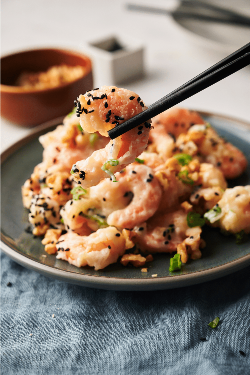 Chop sticks holding a piece of honey walnut shrimp in front of a black plate that is filled with the shrimp.