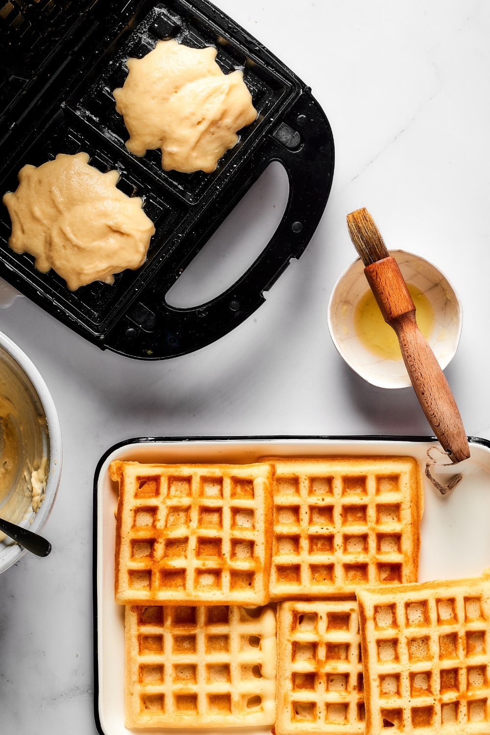A waffle iron with two scoops of batter in it. In front of that is part of a plate with waffles on it.