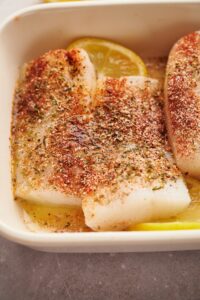 Seasoning on top of two mahi mahi fillets in a baking dish with lemon wedges.