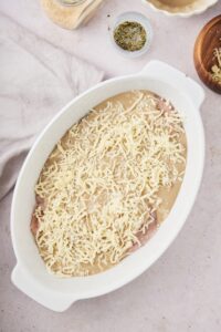 Shredded mozzarella cheese on top of cream of mushroom soup on chicken in a white baking dish.
