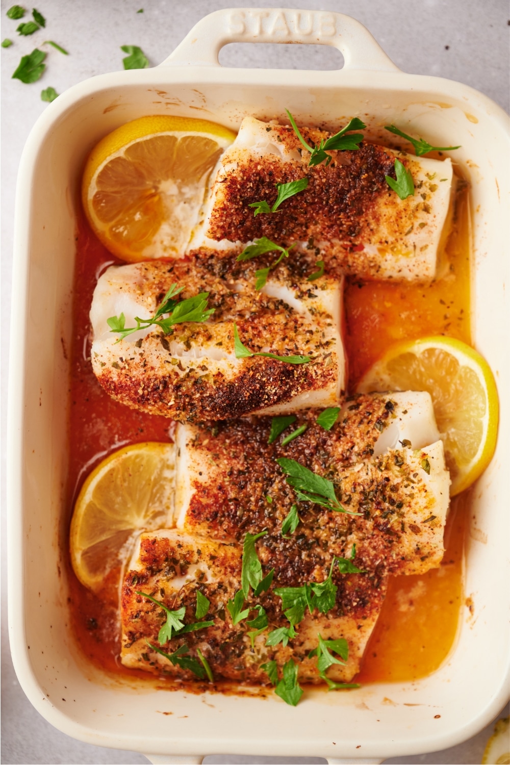 Seasoning on top of four mahi mahi fillets in a white baking dish that has three lemon slices. The dish is on a white counter.