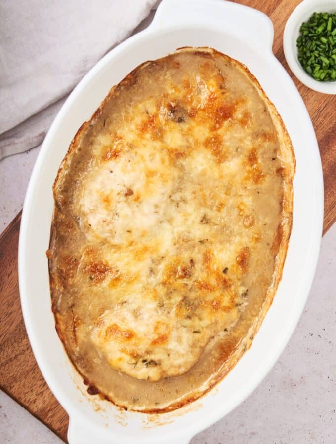 Melted browned cheese on top of chicken breasts in a white baking dish on top of a wooden cutting board.