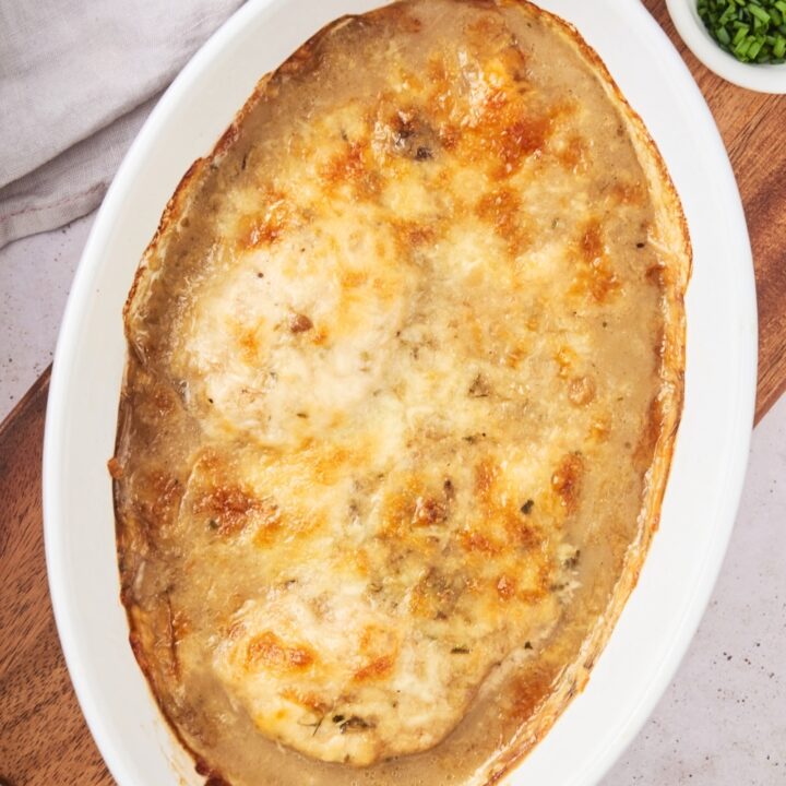 Melted browned cheese on top of chicken breasts in a white baking dish on top of a wooden cutting board.