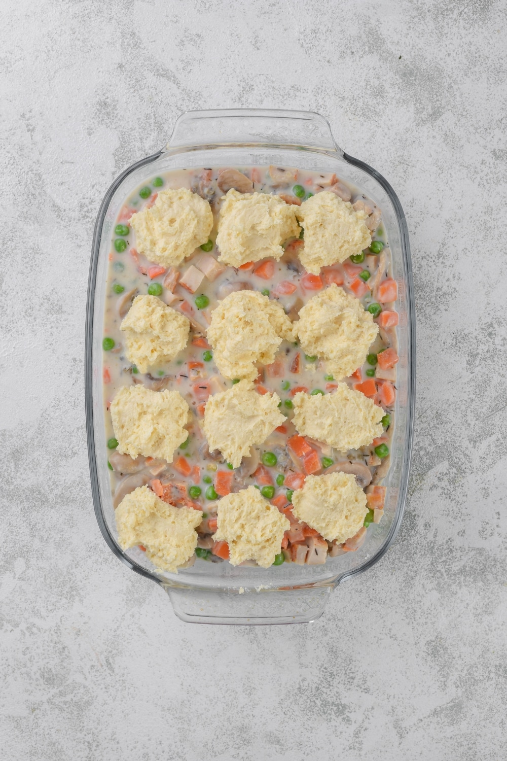 A glass casserole dish full of chicken filling with twelve uncooked drop biscuits on top.