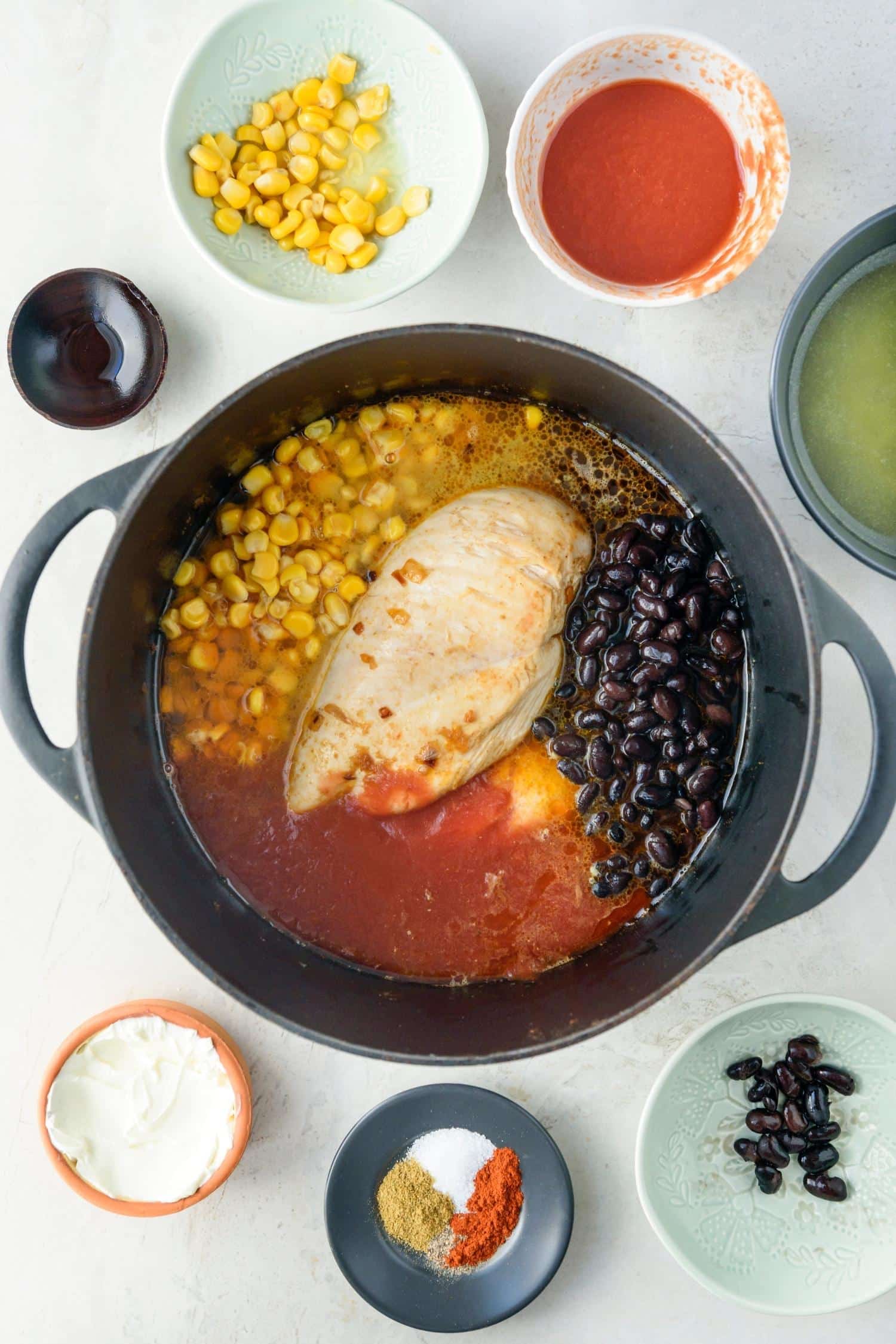 Cooked chicken breast, corn kernels, black beans, tomato sauce, and chicken pot in a large pot, surrounded by bowls of extra ingredients.