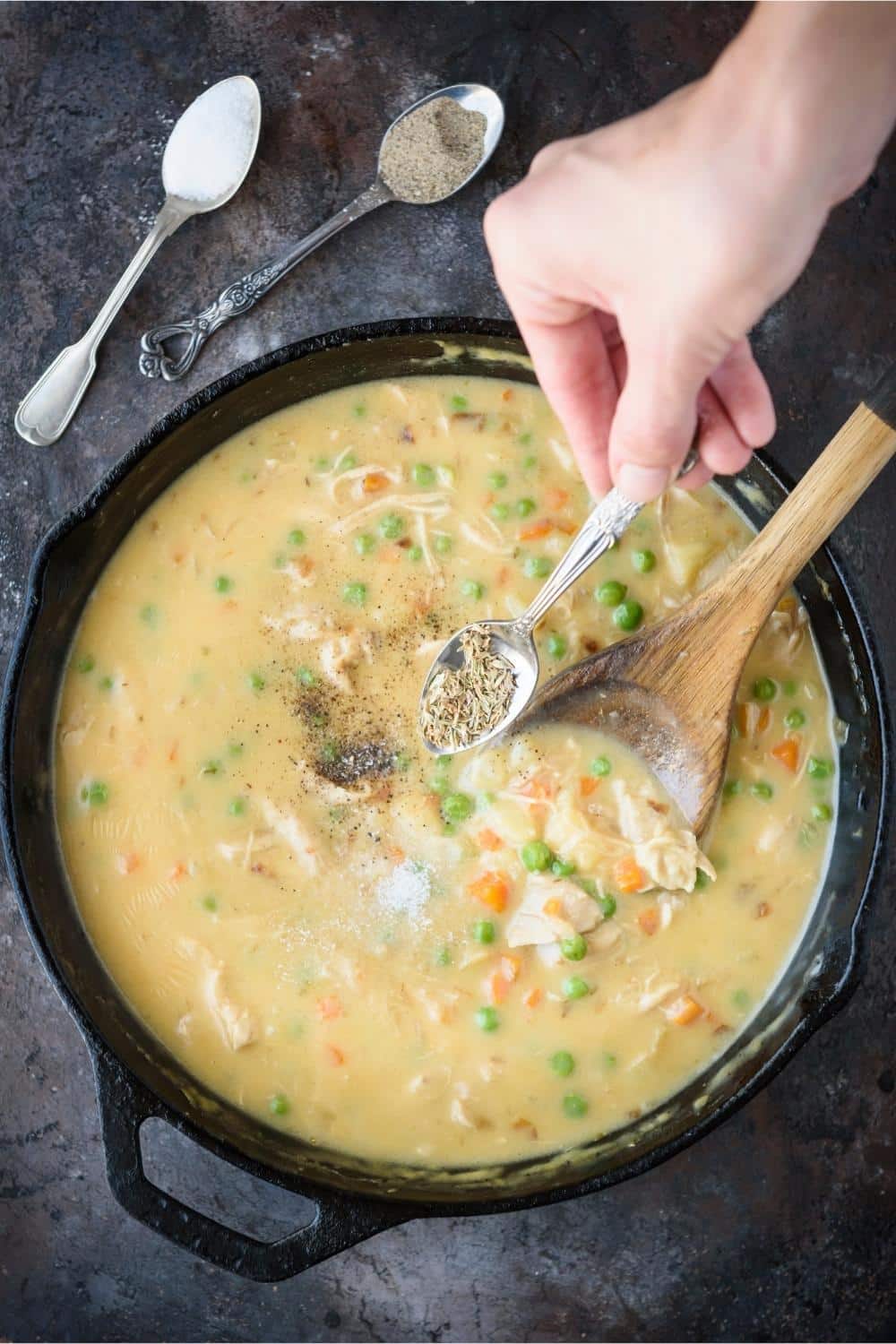 A teaspoon of dried thyme being sprinkled over a cast iron pot of crustless chicken pot pie, next to teaspoonfuls of salt and pepper.