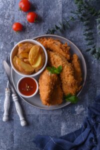 A bunch of chicken tenders on a plate with a cup of potato wedges next to it.