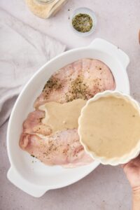 A hand pouring a cup of cream of mushroom soup into a white baking dish that is filled with seasoned chicken.