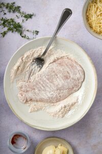 A coated chicken cutlet on a plate of seasoned flour with a fork.