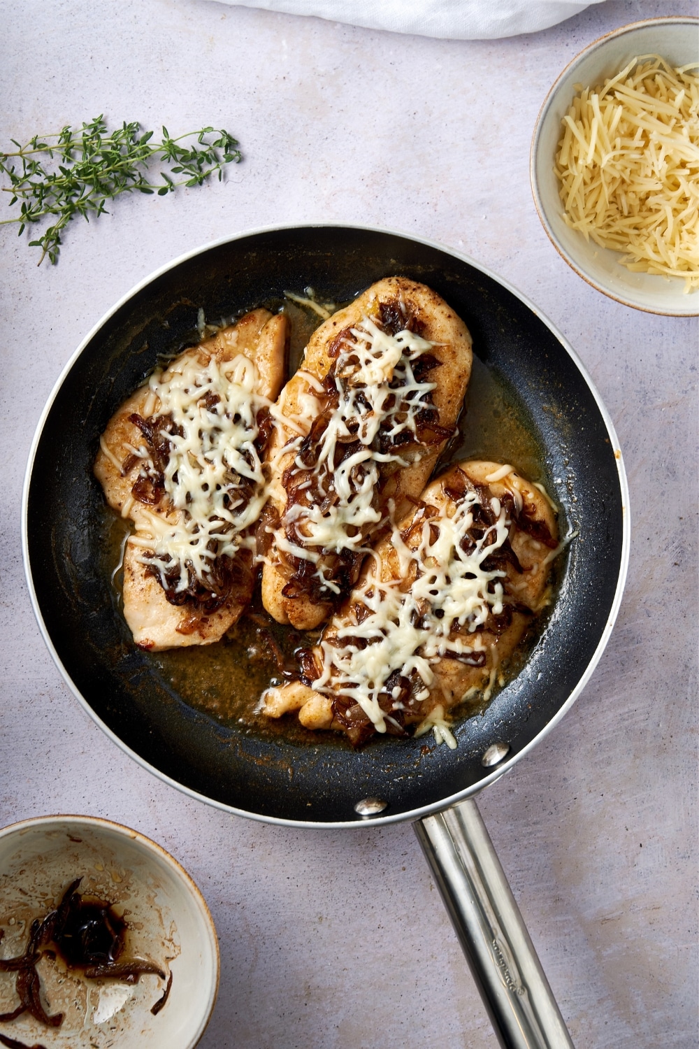 A skillet of chicken cutlets in french onion sauce topped with caramelized onions and melted shredded cheese.