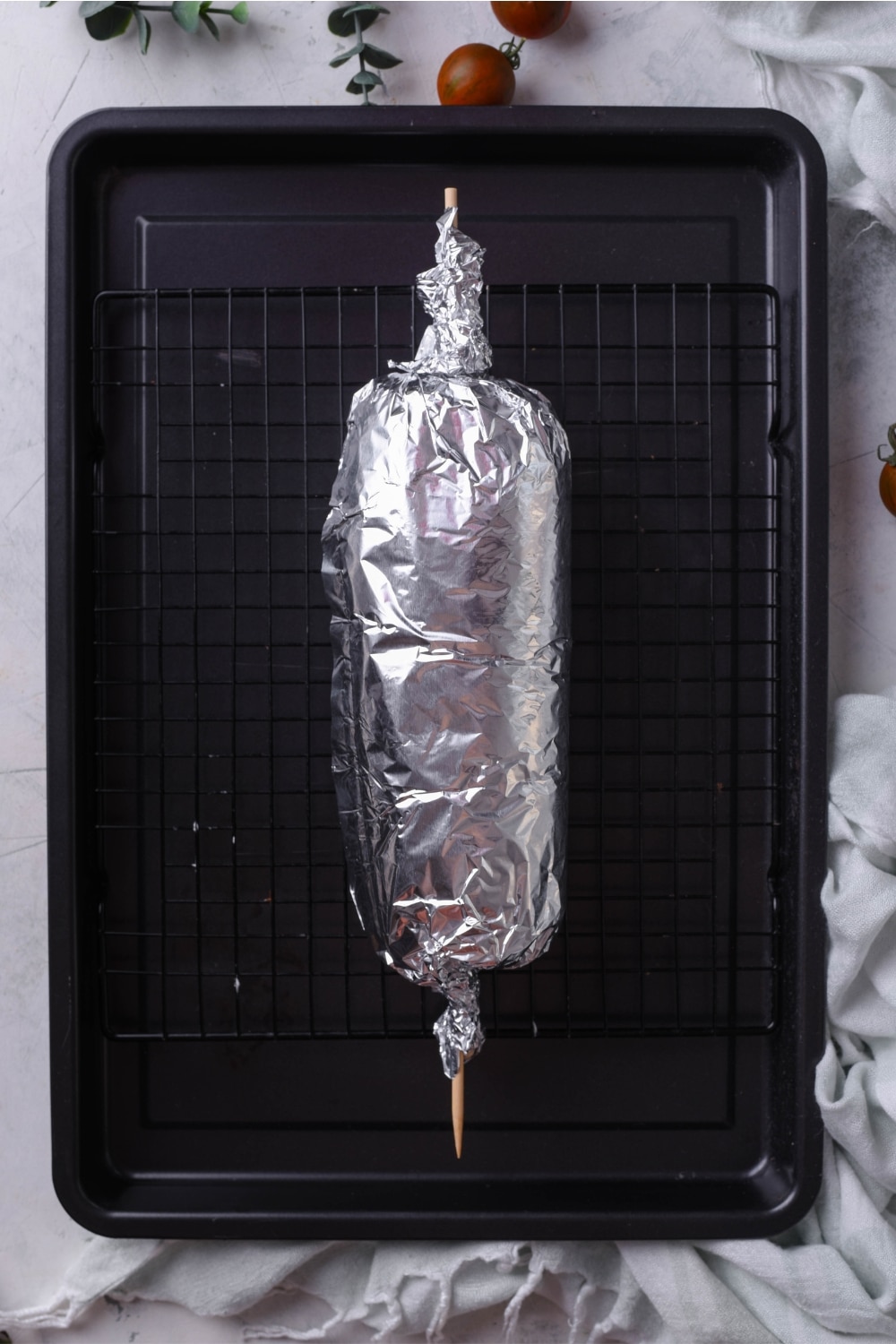 A log of gyro meat wrapped in foil with a skewer down the middle. The meat is on a cooling rack above a baking sheet.