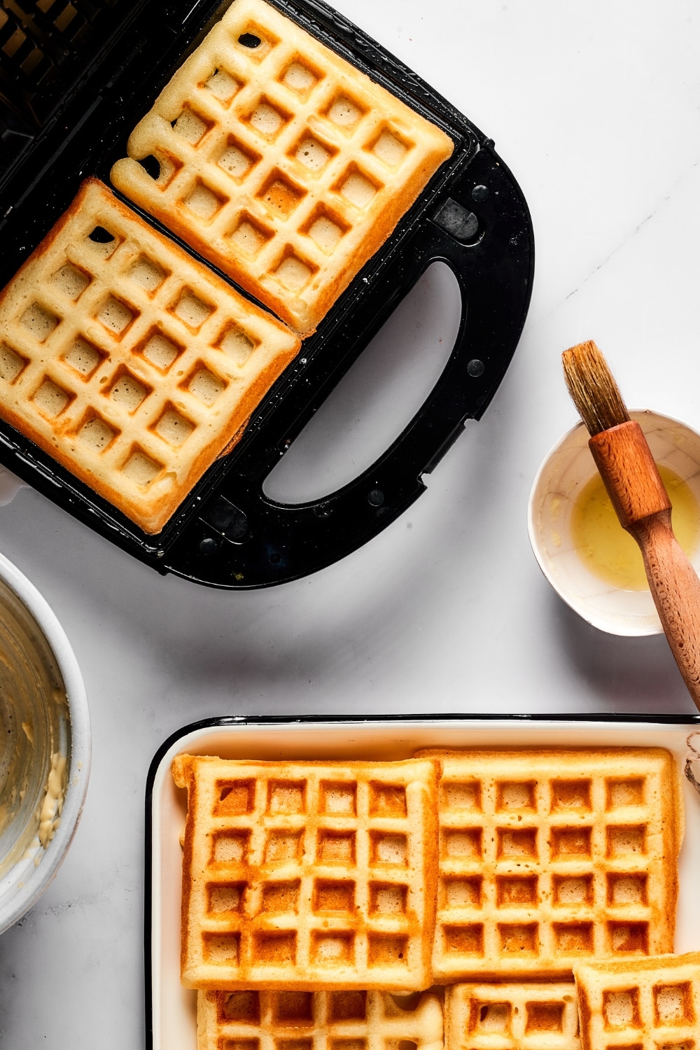 Two cooked waffles in a waffle iron. There are some more waffles in front of it on a plate.