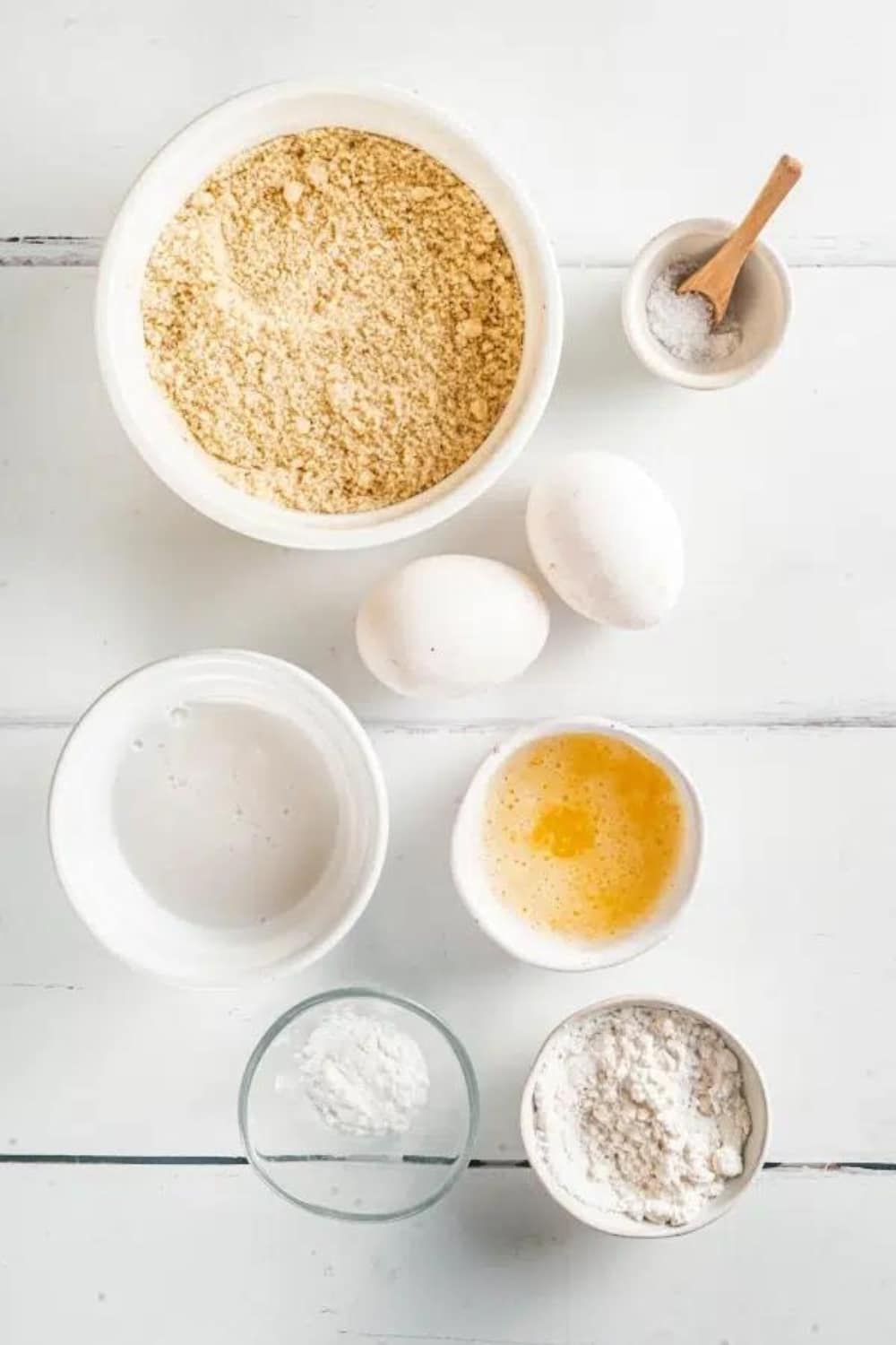 A bowl of almond flour, two eggs, a bowl of milk, a bowl of melted butter, a bowl of sugar, and a bowl of baking soda on a white counter.