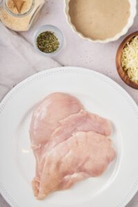 Three pieces of raw chicken overlapping on a white plate. Part of a bowl of cheese, a bowl of seasoning, and part of a bowl of cream of mushroom soup.