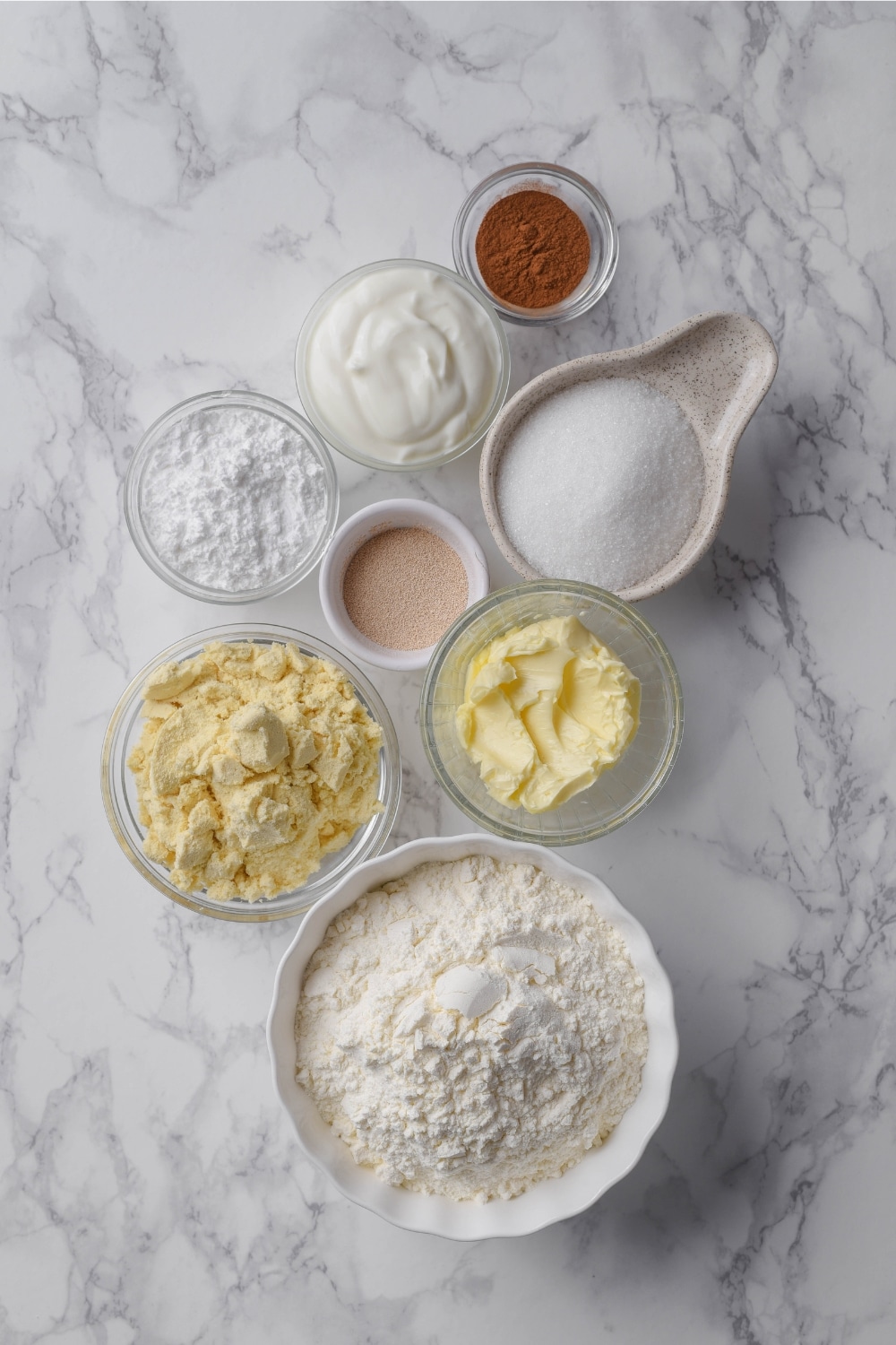 Bowls of flour, protein powder, butter, powdered sugar, white sugar, cinnamon, Greek yogurt, and yeast.