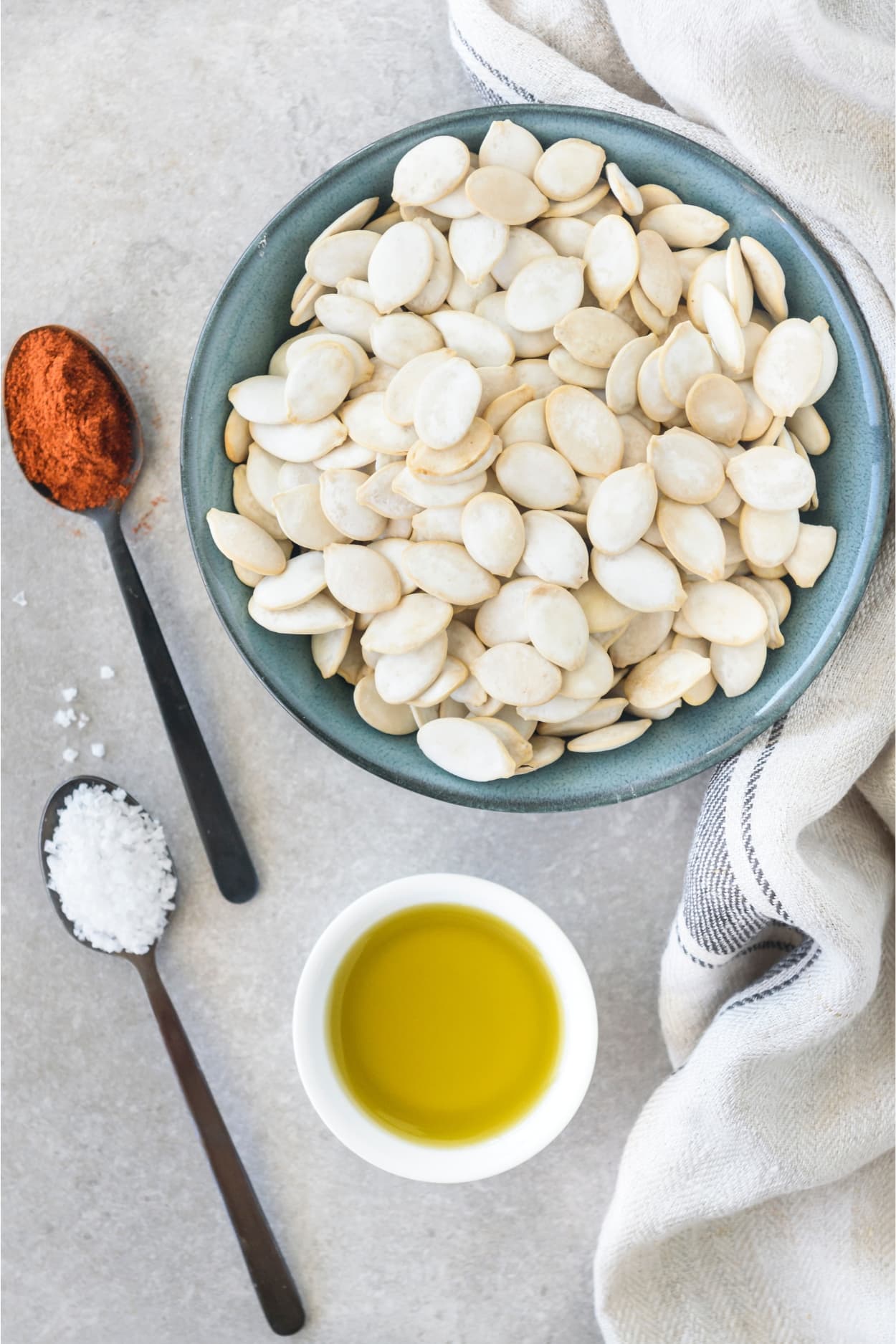 Pumpkin seeds in a bowl, a spoon with cayenne pepper, a spoon with salt, and a bowl of oil all on a white counter.