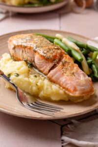 A fillet of salmon on top of mashed potatoes on a white plate with a fork next to the salmon.