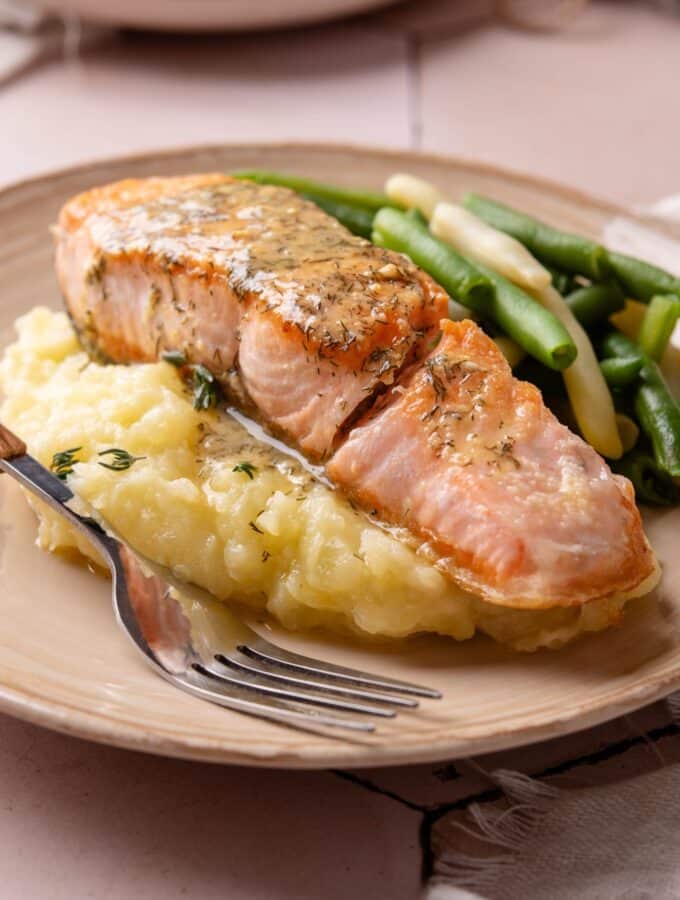 A fillet of salmon on top of mashed potatoes on a white plate with a fork next to the salmon.