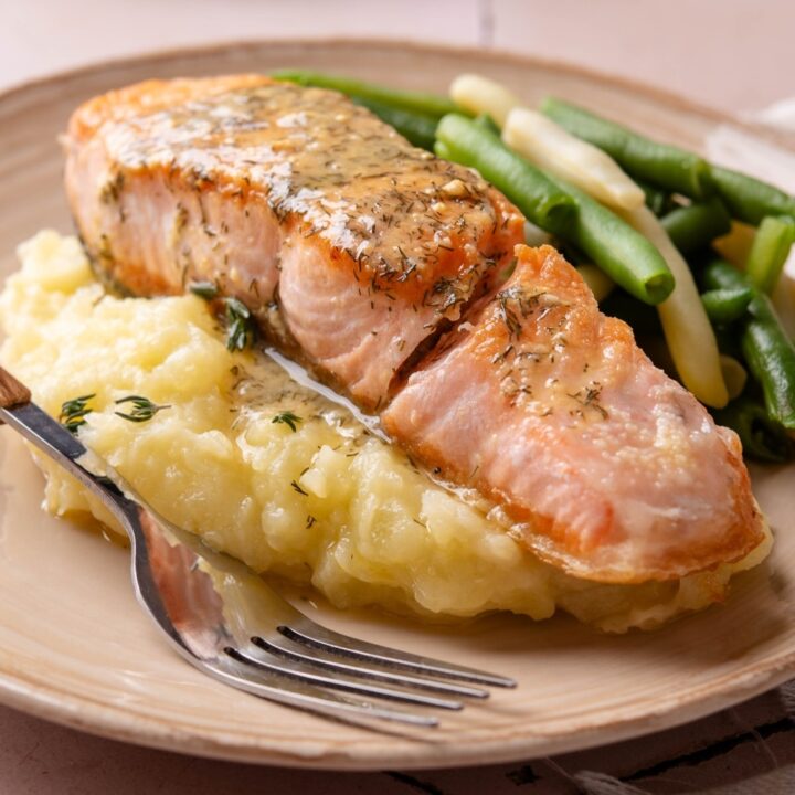 A fillet of salmon on top of mashed potatoes on a white plate with a fork next to the salmon.