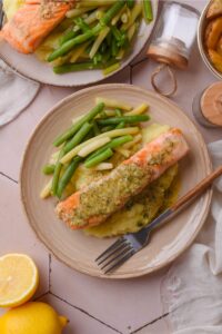 A lemon garlic dill sauce on mashed potatoes on top of a plate. Next to the salmon are green beans.