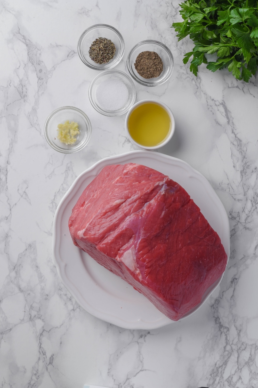 Raw sirloin top on a plate, fresh parsley, and small bowls of olive oil, salt, black pepper, minced garlic, and dried thyme.