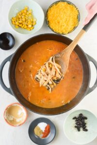 A portion of shredded chicken breast, corn, and black beans on a wooden spatula over a pot of chicken enchilada soup.