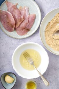 A plate of turkey breast cutlets next to shallow bowls of egg whites and seasoned breadcrumbs and smaller bowls of butter and olive oil.