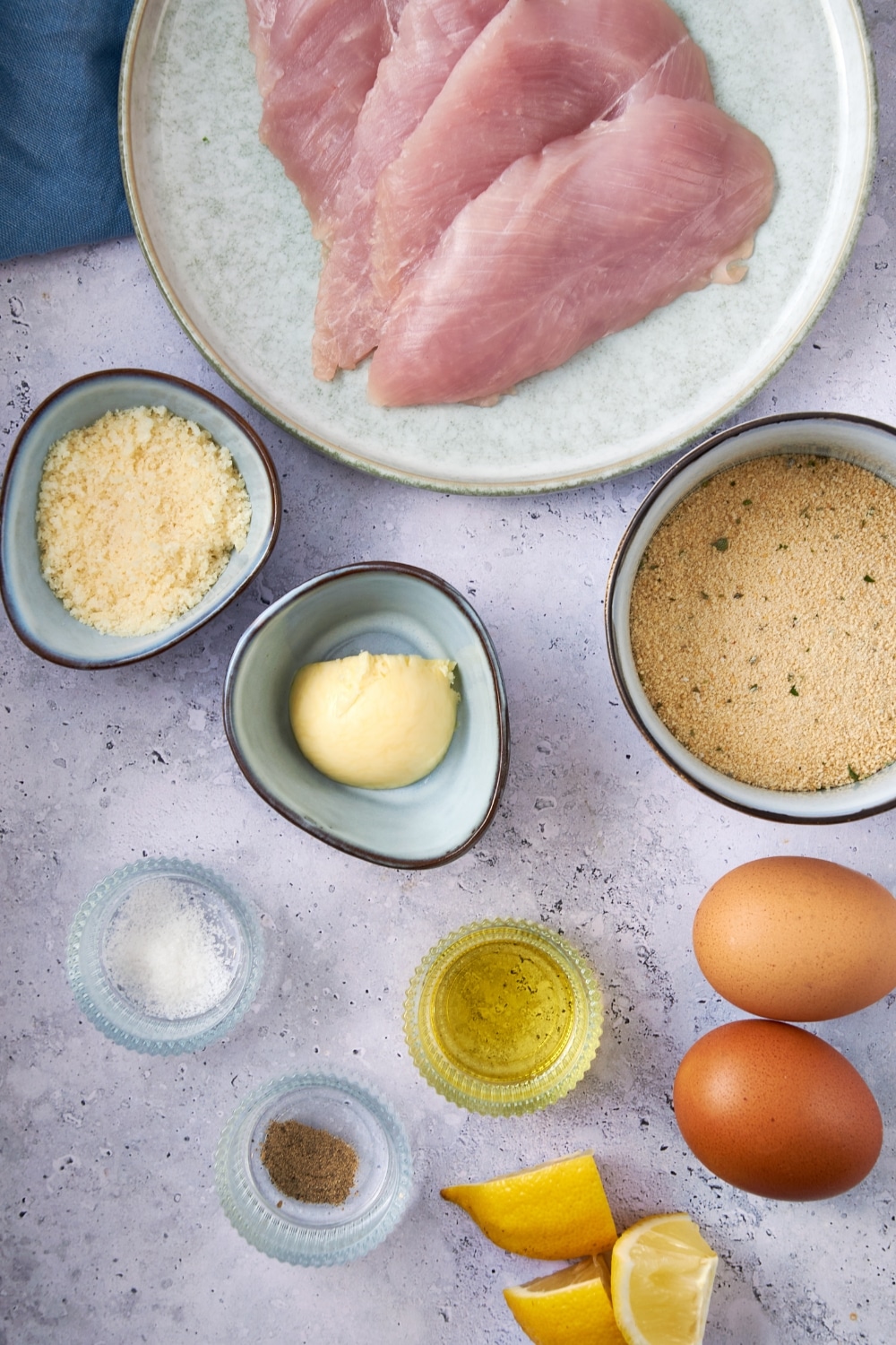 A plate of turkey breast cutlets, bowls of seasoned breadcrumbs, grated parmesan cheese, butter, olive oil, salt, and pepper, eggs, and lemon wedges.