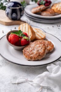 A breakfast plate of two turkey sausages, two toasted baguette slices, and a small bowl of strawberries garnished with mint. A second breakfast plate is seen in the back.