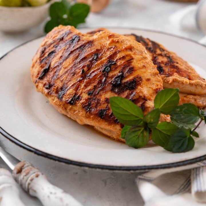 Grilled yogurt-marinated chicken breasts on a white plate, garnished with fresh herbs.