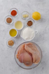 Chicken breasts in a glass bowl, a lemon, and smaller bowls of yogurt, olive oil, lemon juice, minced garlic, and spices.