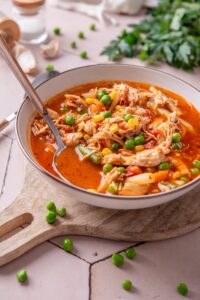 A bowl of soup with tomatoes, shredded chicken, peas, corns, and carrots. There is a spoon in the bowl.