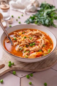 A bowl of soup with tomatoes, shredded chicken, peas, corns, and carrots. There is a spoon in the bowl.