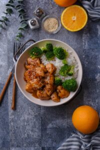 Overhead view of a bowl of orange chicken garnished with sesame seeds served with sides of broccoli and white rice.